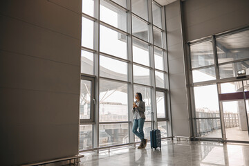 Caucasian woman is standing alone near airport terminal panoramic windows with luggage