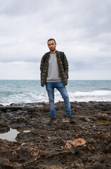 Man posing in the sea with attitude and a backpack. In a sea of rocks surrounded by puddles on a cloudy day. He is wearing a backpack, jeans, sweater and green jacket. His hands are in his pockets.