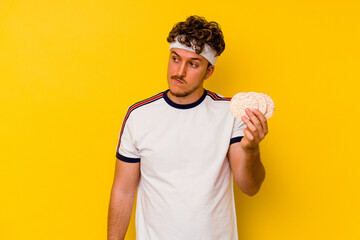 Young sport caucasian man eating a rice cake isolated on yellow background confused, feels doubtful and unsure.