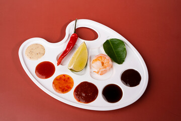 On a red background there is a white palette in which sauces are poured instead of paint: cheese, garlic, barbecue, sweet and sour. There are a slice of lime, a lime leaf, a shrimp, a chili pepper.