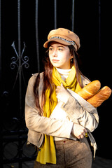 A young fashionable woman sits in a cafe.She's wearing a cap, a beige suit, and a yellow scarf.Baguettes in hand.