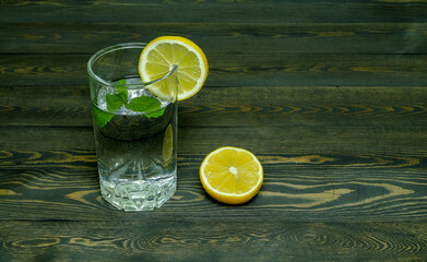 transparent glass glass with water, a slice of lemon and a sprig of mint, and a half of lemon on a dark wooden board table