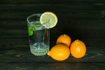 three whole lemons and segment of a lemon on the edge of a transparent glass glass with water and a branch of mint on a dark wooden table from boards