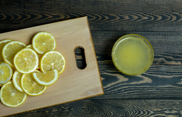 slices of fresh juicy bright yellow lemons laid out on a light wooden cutting board and a glass plate with lemon juice on a dark wooden table, taken close-up