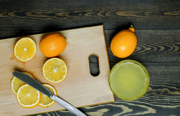 whole lemons and slices of lemons with a knife on a wooden cutting board and next to a small cup with lemon juice on a dark wooden table made of boards