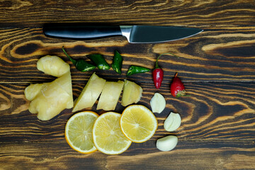 fresh juicy ginger cut into slices, lemon slices, garlic slices, green and red hot peppers and a knife on a dark, coarse wooden background