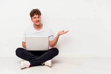 Young caucasian man sitting on the floor holding on laptop isolated on white background showing a copy space on a palm and holding another hand on waist.