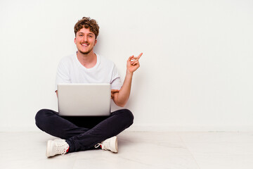 Young caucasian man sitting on the floor holding on laptop isolated on white background smiling cheerfully pointing with forefinger away.