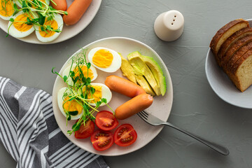 Delicious breakfast, boiled eggs with sausages, tomatoes and avocado, microgreens of peas and sunflowers, plates with food and a bowl of bread on a gray background , top view