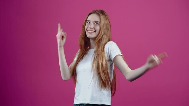 The smiling charismatic young woman monkeys around the room, shows her tongue, rock signs and performs modest dance moves. Freckled teenage girl in a white T-shirt, isolated on a pink background. The