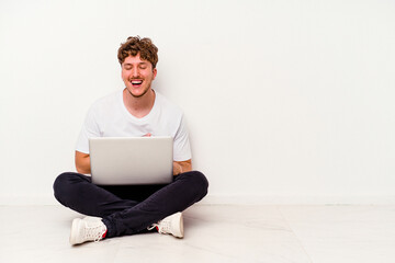 Young caucasian man sitting on the floor holding on laptop isolated on white background laughs happily and has fun keeping hands on stomach.