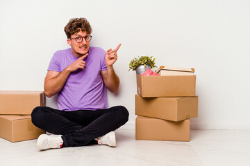 Young caucasian man sitting on the floor ready for moving isolated on white background shocked pointing with index fingers to a copy space.