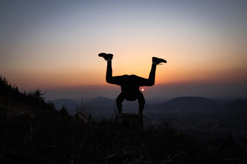 Silhouette of teenager and athlete trying a handstand at sunset in european mountains. Training in natural conditions. Self weight training. Coordination, flexibility.