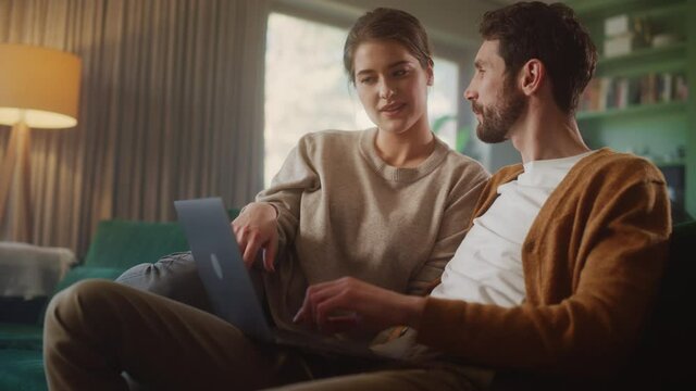 Couple Use Laptop Computer, while Sitting on a Couch in the Apartment. Boyfriend and Girlfriend Talk, Shop on Internet, Choose Product to Order Online, Watch Funny Videos on Streaming Service