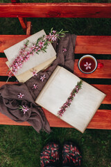 On the bench is an open book with empty pages, a stack of books and almond tree flowers. Next to it is a red cup of tea with a flower. On the green grass, you can see shoes with a design. Read a book 