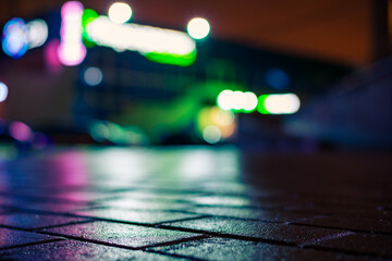 Fototapeta premium Rainy night in the city. Deserted parking mall. Reflections of shop windows on the wet pavement. Colorful colors. Close up view from the pavement level.