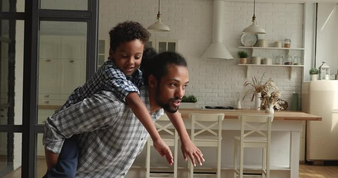 Playful young african father giving piggyback ride to laughing cute kid son at home, cheerful daddy holding on back small child boy, running indoors, mixed race family domestic activity concept.