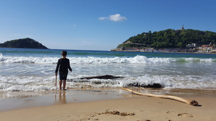 Niño en la orilla de la playa de San Sebastián jugando con troncos traídos por la marea y de fondo los lados de la bahía