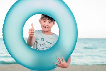 Boy with an inflatable swimming ring on the seashore. Portrait of a boy in the center of a blue swimming circle on the beach. Child dreams of sea travel. Boy shows sign like, thumb up. Travel dreams