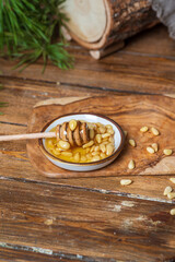 A plate with honey and pine nuts, a honey spoon with transparent honey dripping from it. Light wooden table background, behind a pine branch.