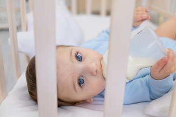 baby boy eating from milk bottle in crib in blue bodysuit, cute little baby in bedroom, baby food concept
