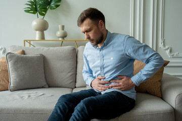 A man suffers from abdominal pain while sitting at home on the couch. Beautiful young man suffering...
