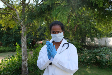 Young Indian female wearing a doctor uniform, gloves, and stethoscope praying in the park