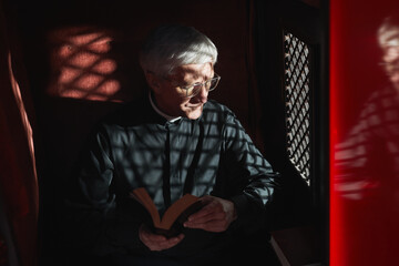 Senior priest sitting with the Bible in confessional and listening to confession - obrazy, fototapety, plakaty