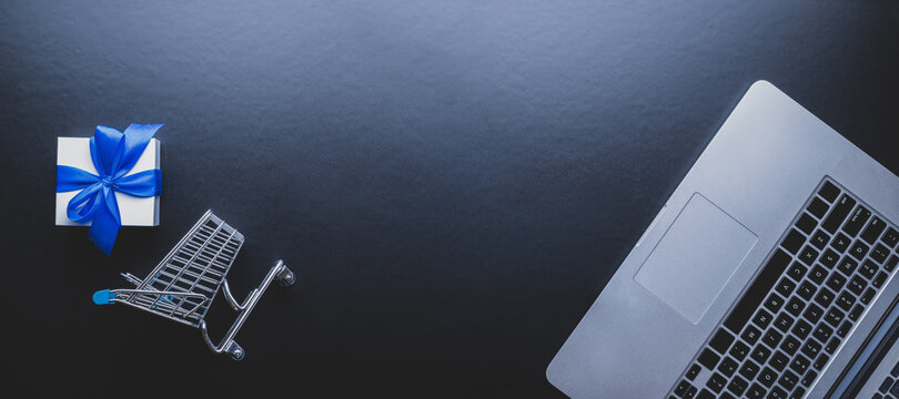 Ecommerce. Laptop computer, shopping trolley and white gift with blue ribbon on dark background. Website retail business concept. Easy Ecommerce Website Shop.