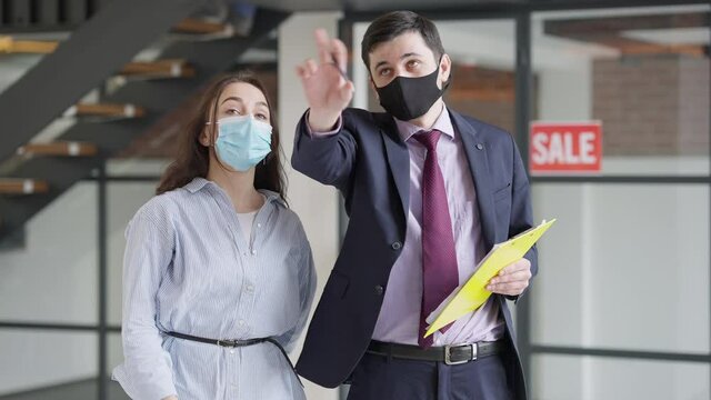 Client and broker in coronavirus face masks talking gesturing and shaking hands standing in new premises indoors. Portrait of confident Caucasian woman and man in new flat on Covid pandemic