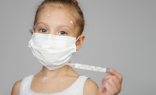 Portrait Of Sick Little Child Girl With Chickenpox In Protective Face Mask.  Toddler With Varicella Virus Or Chickenpox Bubble Rash With Fever. Hold A Thermometer In Hand