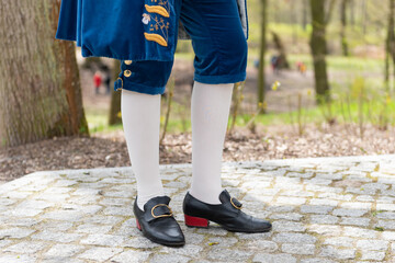 Details of a man's dress in a baroque costume. White stockings and black shoes, golden buttons, decorative hems. A man standing on a cobbled walkway in the background of a blurred garden backdrop. - 430242388