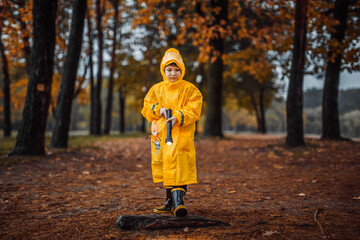A little boy in a yellow raincoat and rubber boots plays on the street with a flashlight in an autumn park in yellow leaves. Portrait of a little happy and cheerful preschooler