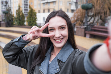 Beautiful brunette girl makes a selfie on a smartphone and shows the gesture of peace while walking on the street