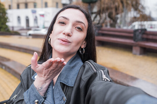 Beautiful brunette girl makes a selfie and shows a kiss gesture on a smartphone while walking on the street