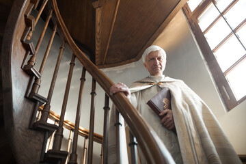 Portrait of senior priest holding Bible and looking at camera while standing on wooden stairs in...