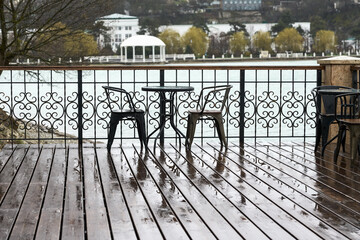 Summer cafe on the lake with mountain views. Behind the wrought-iron fence on the wooden deck is a...