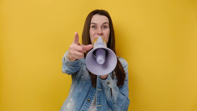 Portrait of shocked young girl screaming in megaphone spreading hands point finger camera on you, dressed in denim jacket, posing isolated on yellow background wall. People emotion lifestyle concept