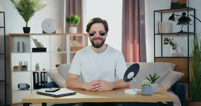 Handsome smiling happy caring young bearded guy poses at camera with anti-aging hydrogel eye-patches sitting at the table in cozy room,slow motion