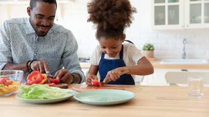 Happy African American daughter kid helping dad to prepare dinner, slicing vegetables, fresh pepper...