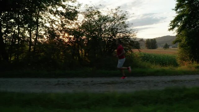 Wide shot of a Caucasian man in sportswear running along a dirt road near cornfields
