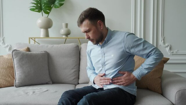 A man suffers from abdominal pain while sitting at home on the couch. Beautiful young man suffering from abdominal pain sitting on the couch at home