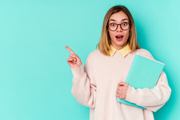 Young student woman holding books isolated on blue background pointing to the side