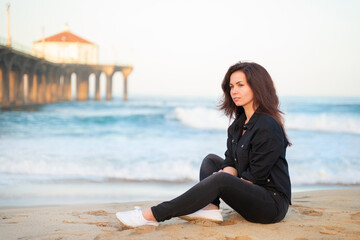 Fototapeta na wymiar A smiling young woman with long hair sits on the sand in front of a pier on Manhattan Beach in Los Angeles in the early morning