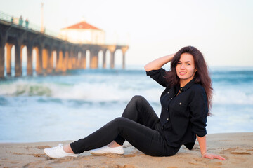 A smiling young woman with long hair sits on the sand in front of a pier on Manhattan Beach in Los Angeles in the early morning