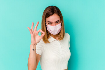 Young woman wearing a mask for virus isolated on blue background cheerful and confident showing ok...