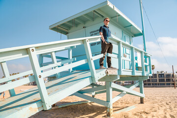 Handsome young blonde man in shirt poses on light blue lifeguard tower on Los Angeles beach
