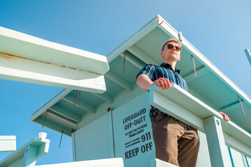 Handsome young blonde man in shirt poses on light blue lifeguard tower on Los Angeles beach
