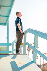 Handsome young blonde man in shirt poses on light blue lifeguard tower on Los Angeles beach