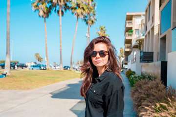 Cute young brunette girl in sunglasses walks along the Los Angeles waterfront with palm trees and streets on a summer day
